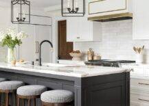 Backless stools sit at a black center island topped with a statuary marble countertop holding a sink with a matte black gooseneck faucet lit by two black gothic lanterns.