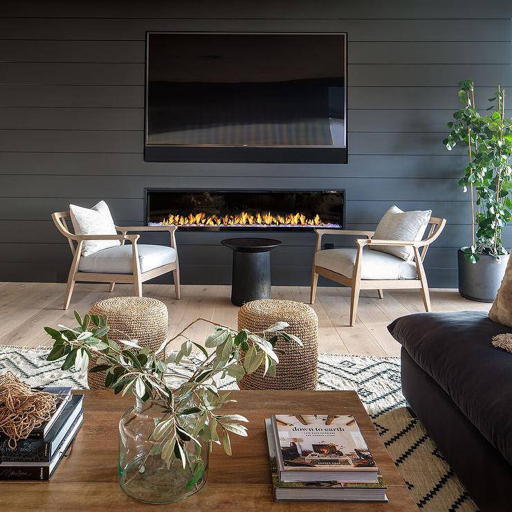 Living room features a black shiplap fireplace under a flatscreen TV, beige vintage accent chairs with a black pedestal accent table and woven stools at a wooden coffee table.