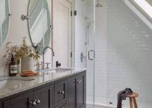 Two vintage barn sconces light oval pivot mirrors hung above a black wooden dual washstand fitted with a slatted shelf and donning polished nickel ball knobs and a gray marble countertop finished with polished nickel gooseneck faucets.
