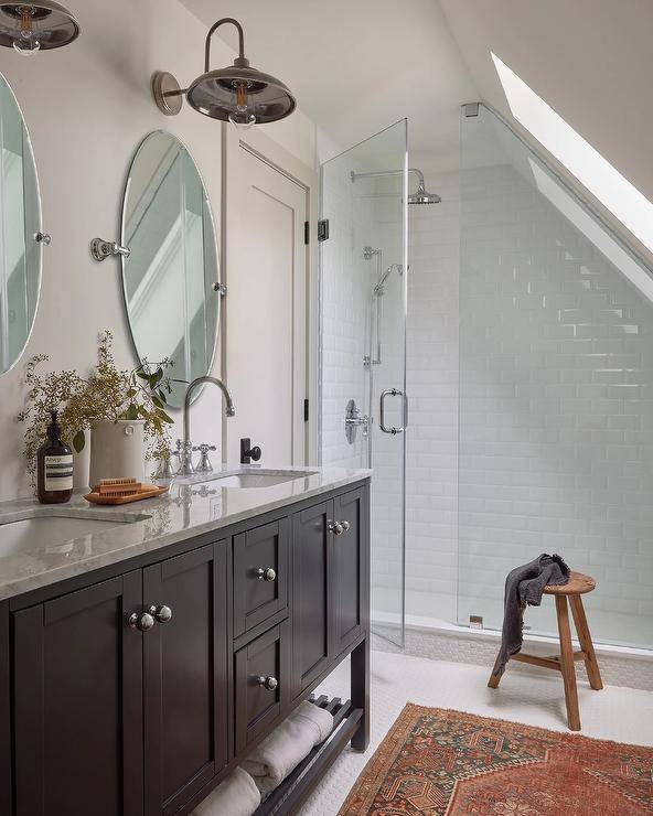 Two vintage barn sconces light oval pivot mirrors hung above a black wooden dual washstand fitted with a slatted shelf and donning polished nickel ball knobs and a gray marble countertop finished with polished nickel gooseneck faucets.