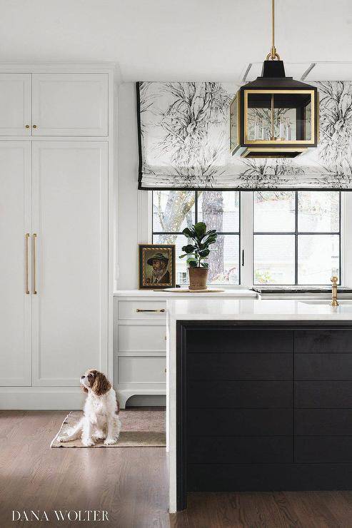 Chic gold and black kitchen features floor-to-ceiling white cabinets donning brass hardware and fixed beside a row of windows covered in a black and white floral roman shade. A black island is contrasted with a white marble waterfall countertop lit by black and gold French lanterns.