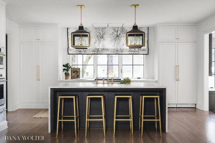 Gold and black French lanterns illuminate a black kitchen island contrasted with a white marble waterfall edge countertop finished with a sink and brass faucet kit. Gold and black stools sit at the island and complement this white, gold, and black kitchen. A white and black floral roman shade with black trim hangs from a row of windows located above and beside white cabinets and a white wood paneled refrigerator.