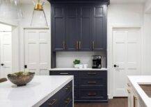 Kitchen features black cabinetry adorned with brass pulls over a white quartz slab backsplash, and a white topped island with black drawers and brass pulls.