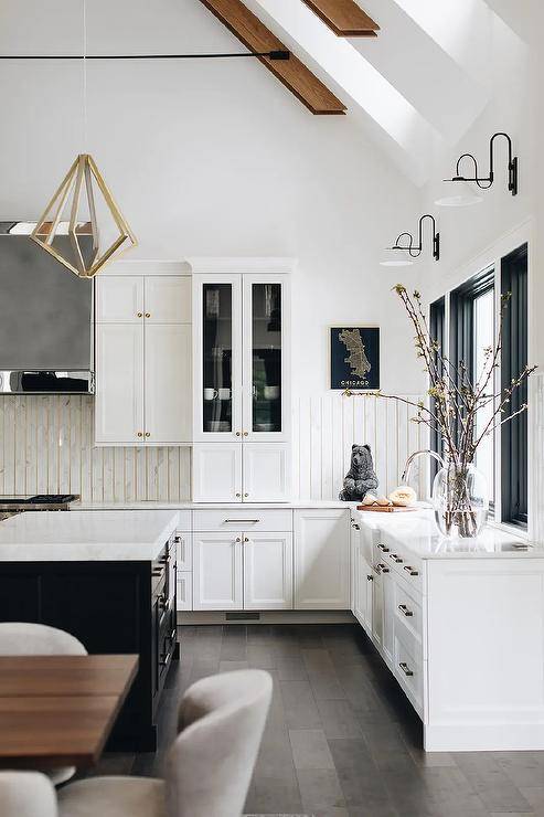 White and black kitchen features a brass geometric lantern hung over a black island topped with a marble countertop and framed by a dark stained wood floor. The island is contrasted with white perimeter cabinets, while white and black vintage sconces light black framed window positioned above a sink with a polished nickel gooseneck faucet.