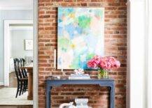 Modern foyer features exposed brick walls, pink green and blue abstract art over a dark gray chinoiserie console table with an elephant accent table stool atop wooden floors.