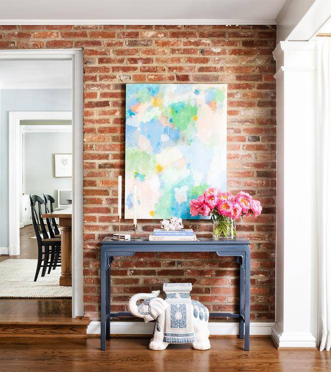 Modern foyer features exposed brick walls, pink green and blue abstract art over a dark gray chinoiserie console table with an elephant accent table stool atop wooden floors.