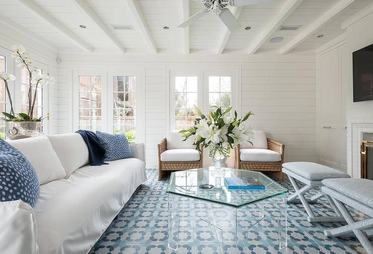 Welcoming white and blue living room boasts a lucite hexagon coffee table placed on white and blue mosaic floor tiles in front of a white slipcovered sofa accented with blue pillows. A white ceiling fan is mounted to a white plank ceiling, while light blue x-stools and curved rope and rattan accent chairs are placed at the coffee table.