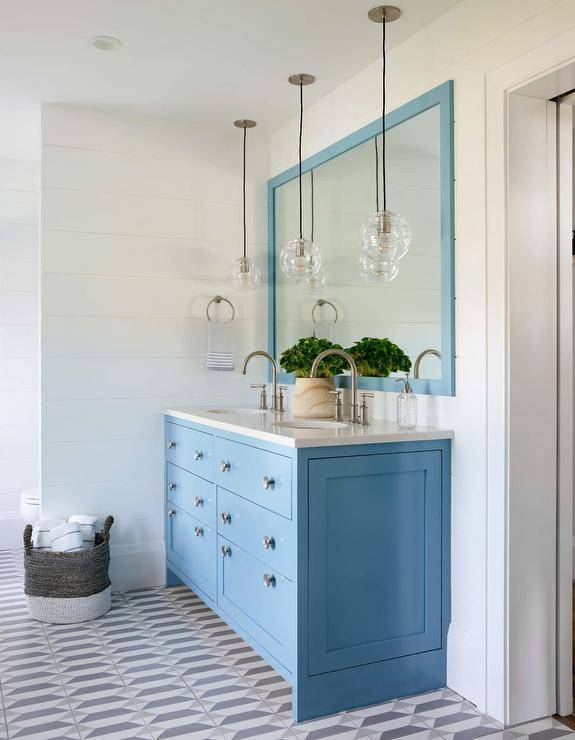 Placed on white and gray geometric floor tiles, a blue double bath vanity is adorned with satin nickel knobs and satin nickel gooseneck faucets fixed to a marble countertop. The vanity is illuminated by staggered glass globe lights hung in front of a blue framed vanity mirror mounted against white shiplap trim.