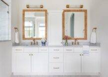 Serena & Lily Balboa Rattan Mirrors hang in a gorgeous bathroom over a white dual bath vanity adorned with brass pulls and aged brass hook and spout faucets fixed to a marble countertop.