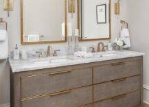 Brass sconces light brass rivet mirrors hung over a gray shagreen double washstand fitted with brass hardware and brass faucets mounted to a marble countertop.