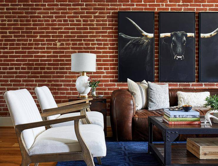 A steer triptych art piece is hung from a red brick wall over a brown leather sofa accented with white and gray pillows in a modern country living room. An industrial rivet coffee table sits in front of the sofa on a bold blue rug and seats two wooden accent chairs with white cushions.