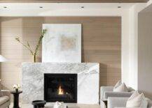 A white and brown abstract art piece sits on a marble beveled fireplace mantel against a light brown plank wall.