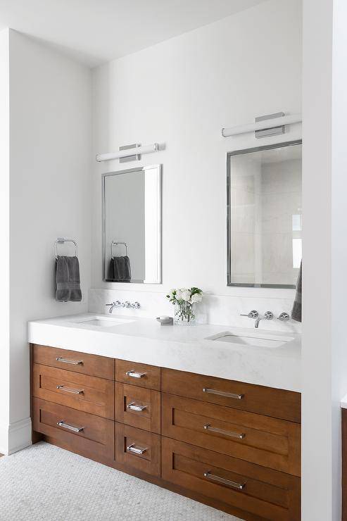 Bathroom features a brown wooden double vanity topped with white marble under nickel mirrors, illuminated by long white glass sconces and light gray floor tiles.