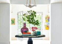 Luxurious entryway features a gold and black octagon table paired with fuchsia stools and placed on a white and blue rug beneath a blue tray ceiling fitted with a white and gold pendant.