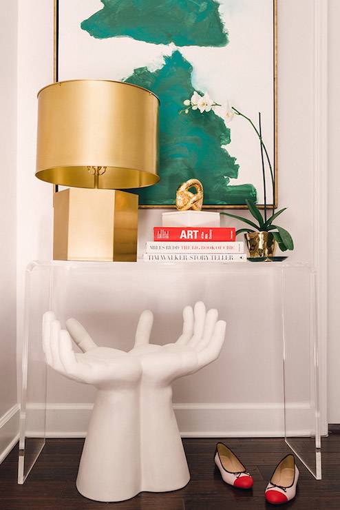 Stylish entryway with CB2 Peekaboo Clear Console Table, with Studio A Hands Stool tucked below, topped with a Mary Mcdonald Pythagoras Table Lamp, stacked books topped with a gold knot, Hillary Thomas Malachite Bowls and a shiny gold planter filled with a white orchid against a backdrop of emerald green abstract art mounted in a gold floating frame.