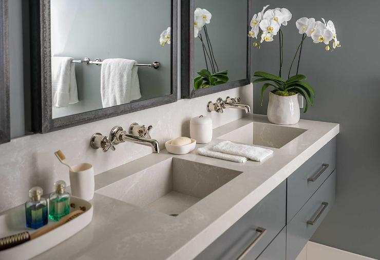 A gray floating dual sink vanity topped with a light gray quartz countertop is accented with polished nickel pulls and polished nickel faucets mounted to a light gray quartz backsplash under galvanized metal framed medicine cabinets fixed to gray walls.