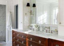 A cherrywood dual washstand accented with vintage hardware is fitted with a honed marble countertop finished with satin nickel hook and spout faucets. A white framed mirror hangs over the washstand and between white linen cabinets.