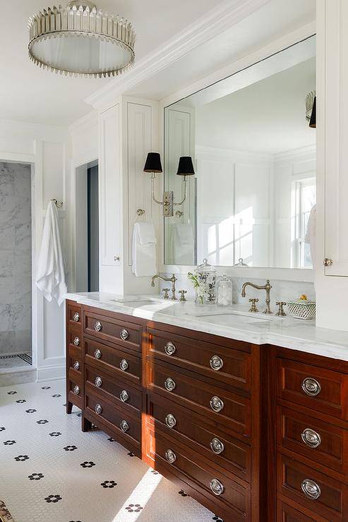 A cherrywood dual washstand accented with vintage hardware is fitted with a honed marble countertop finished with satin nickel hook and spout faucets. A white framed mirror hangs over the washstand and between white linen cabinets.