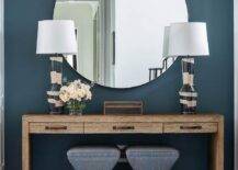 Benjamin Moore Nocturnal Gray pain boasts a bold finish in a foyer entryway styled with a wooden console table, a large round mirror, and charcoal gray stools. A set of gray and black lamps decorate the console table with a contemporary finish.