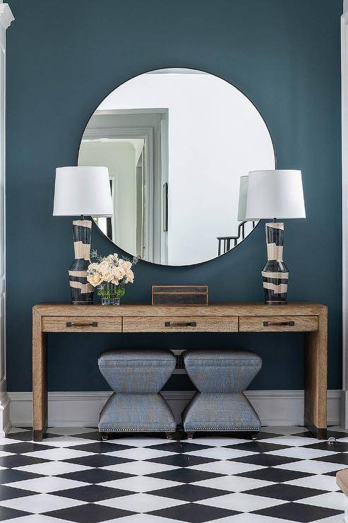 Benjamin Moore Nocturnal Gray pain boasts a bold finish in a foyer entryway styled with a wooden console table, a large round mirror, and charcoal gray stools. A set of gray and black lamps decorate the console table with a contemporary finish.