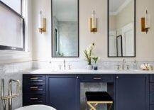 Bathroom features tall black vanity mirrors flanked by brass and white sconces over a gray marble vanity backsplash and a blue double washstand with brass pulls, a gray marble countertop with a sink that boasts polished nickel faucets and a gold and black vanity stool.