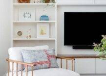 A wall of white styled built-ins surround a flat panel TV mounted against a white wall, as a vintage wooden spindle chair sits on a gray jute rug.