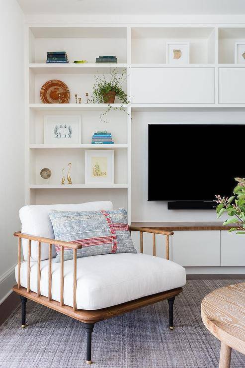A wall of white styled built-ins surround a flat panel TV mounted against a white wall, as a vintage wooden spindle chair sits on a gray jute rug.
