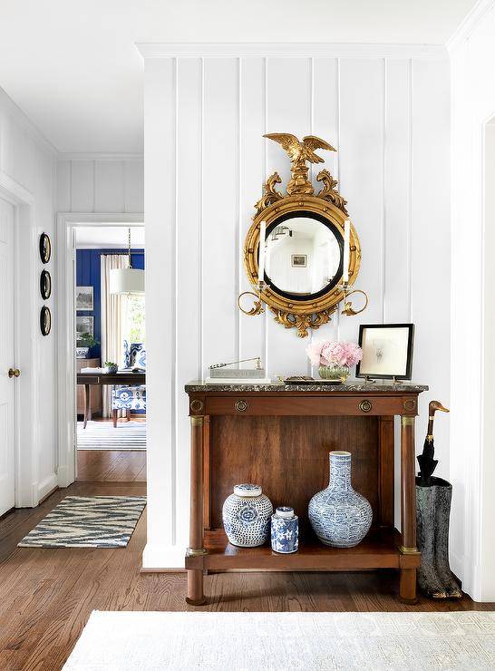 Entryway features an antique console table with black marble countertop boasting Chinese ginger jars, underneath a gold eagle mirror, vertical plank foyer wall and a boots umbrella holder.