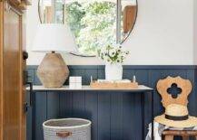 Cottage style foyer features a round black mirror on a white wall over blue vertical shiplap trim and a vintage reeded lamp atop an iron and marble console table with white and gray woven bins.