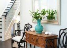 Beautiful foyer with vintage console with drawers topped with seashells and a turquoise urn filled with white lilies in front of a silver leafed mirror with black Chippendale faux bamboo armchairs on either side.