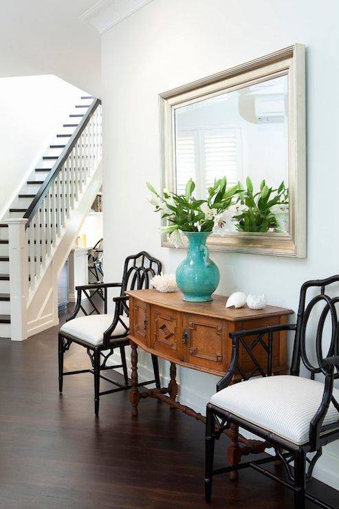 Beautiful foyer with vintage console with drawers topped with seashells and a turquoise urn filled with white lilies in front of a silver leafed mirror with black Chippendale faux bamboo armchairs on either side.