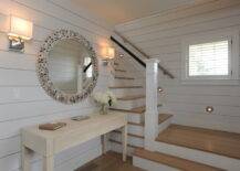 Coastal foyer boasts walls clad in shiplap over a white washed two drawer console table situated below a round oyster shell mirror flanked by a pair of contemporary nickel sconces alongside stairwell with wire spindles lit by round step lights.