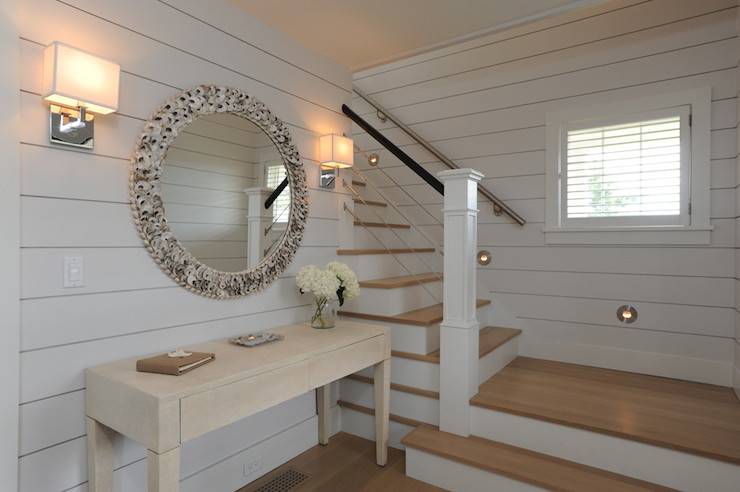 Coastal foyer boasts walls clad in shiplap over a white washed two drawer console table situated below a round oyster shell mirror flanked by a pair of contemporary nickel sconces alongside stairwell with wire spindles lit by round step lights.