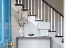 Entryway features a white capiz lamp on a gray waterfall console table in a stairwell, a pink and blue vintage runner, tan woven baskets and a glossy blue front door, lit by a chandelier.