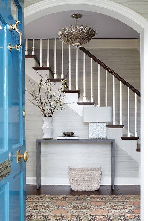 Entryway features a white capiz lamp on a gray waterfall console table in a stairwell, a pink and blue vintage runner, tan woven baskets and a glossy blue front door, lit by a chandelier.