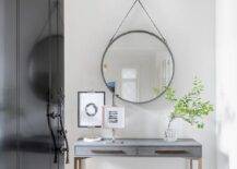 A entryway features a glossy black front door accented with an ornate bronze door handle and a round gray mirror mounted over a gold and gray shagreen console table with a green stool and gray rug.