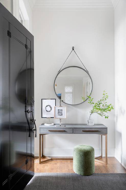 A entryway features a glossy black front door accented with an ornate bronze door handle and a round gray mirror mounted over a gold and gray shagreen console table with a green stool and gray rug.