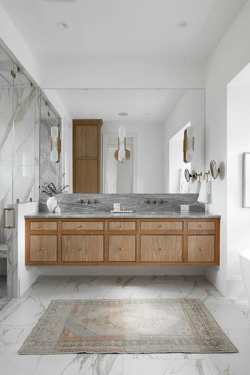 Marble floor tiles are fixed beneath a brown floating dual washstand adorned with brass knobs and a gray marble countertop. A brass faucet kits are fixed to a gray marble backsplash beneath a frameless vanity mirror finished with three brass and alabaster sconces.