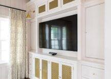 Brass metal grill front TV cabinets accented with brass and glass pulls flank a flat panel TV mounted in a niche between white cabinets and drawers lit by gold conical sconces.