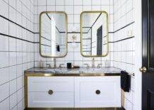 Curved brass vanity mirrors hang from a black and white tiled wall on either side of a brass and white glass 2-light globe scone and over a white dual washstand with brass trim. The washstand, placed on black and white geometric floor tiles, is topped with a gray marble countertop finished with aged brass gooseneck faucets.