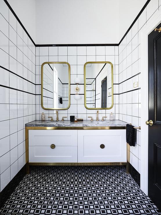 Curved brass vanity mirrors hang from a black and white tiled wall on either side of a brass and white glass 2-light globe scone and over a white dual washstand with brass trim. The washstand, placed on black and white geometric floor tiles, is topped with a gray marble countertop finished with aged brass gooseneck faucets.