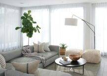 Framed by windows covered in sheer white curtains, this wonderfully designed living room features a fiddle leaf fig placed behind a pebble gray sectional complemented iwth French burlap pillows. The sofa is matched with a round black coffee table placed on a gray rug, while a black swing arm floor lamp with a white drum shade lights two white poufs.