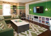 Green family room features a green accent wall lined with a flatscreen TV flanked by pendants above a low built in media shelving unit. A green tufted sectional topped with green accent pillows stands beside a storage coffee table atop a green lattice rug.