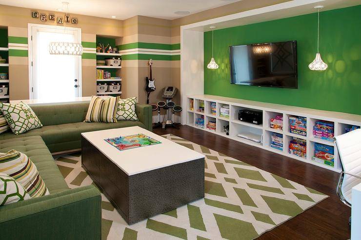 Green family room features a green accent wall lined with a flatscreen TV flanked by pendants above a low built in media shelving unit. A green tufted sectional topped with green accent pillows stands beside a storage coffee table atop a green lattice rug.