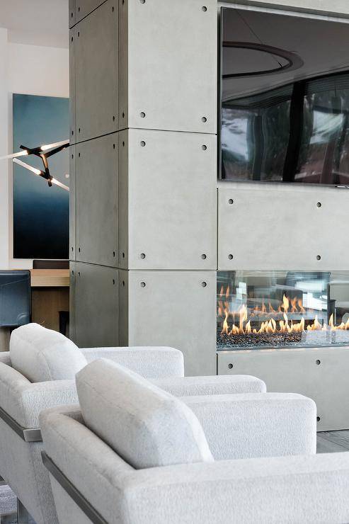A large flat paneled TV is fixed in a niche over a gray industrial style double-sided fireplace warming two side-by-side light gray accent chairs.