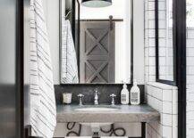 A concrete top washstand accented with oil rubbed bronze legs is fitted with a polished nickel cross handle faucet mounted beneath a tall black framed mirror illuminated by a black industrial pendant.