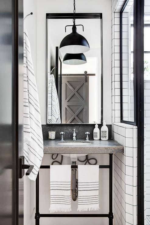 A concrete top washstand accented with oil rubbed bronze legs is fitted with a polished nickel cross handle faucet mounted beneath a tall black framed mirror illuminated by a black industrial pendant.