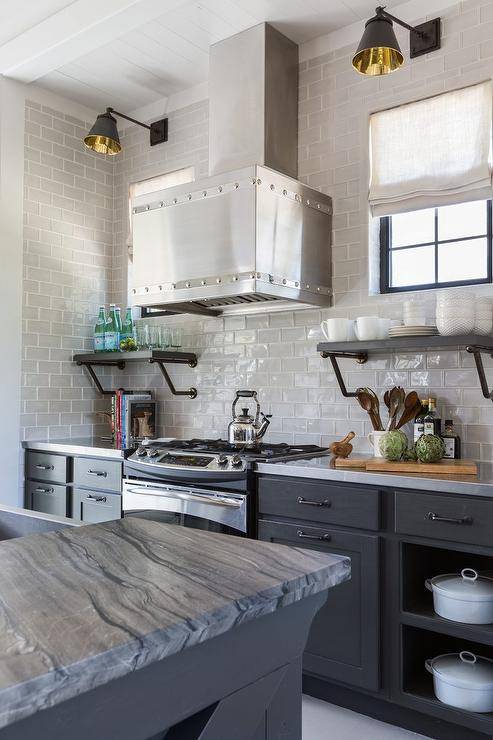 Gray and black kitchen design features a stainless steel industrial hood flanked by black and brass sconces on gray glass subway tiles over a cooktop, black industrial shelves over gray cabinets, and a gray kitchen island with dark gray marble countertop.