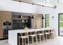 Black industrial pendants hang a rustic wood beam over a black kitchen island finished with a white quartz waterfall countertop. The countertop is finished with an induction cooktop and seats wishbone counter stools.