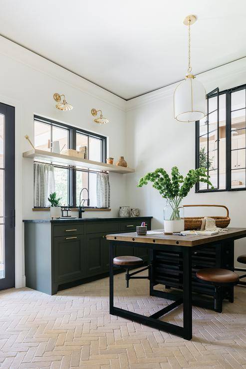 Kitchen features an industrial wood and metal desk with built in leather stools on faded red brick herringbone pavers lit by a white and gold cage lantern and green cabinets.